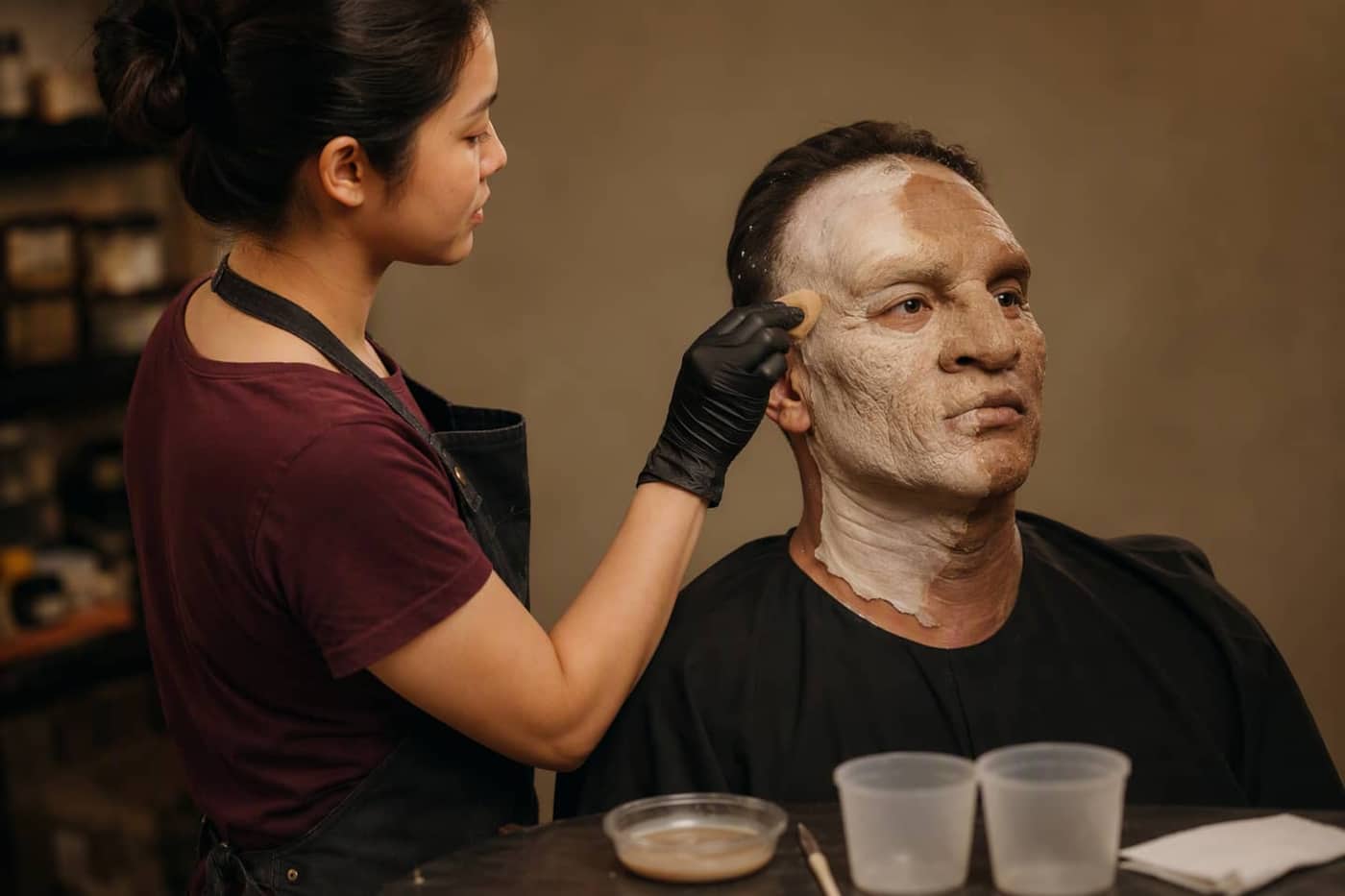Artist painting a full-face silicone character prosthetic on a seated model
